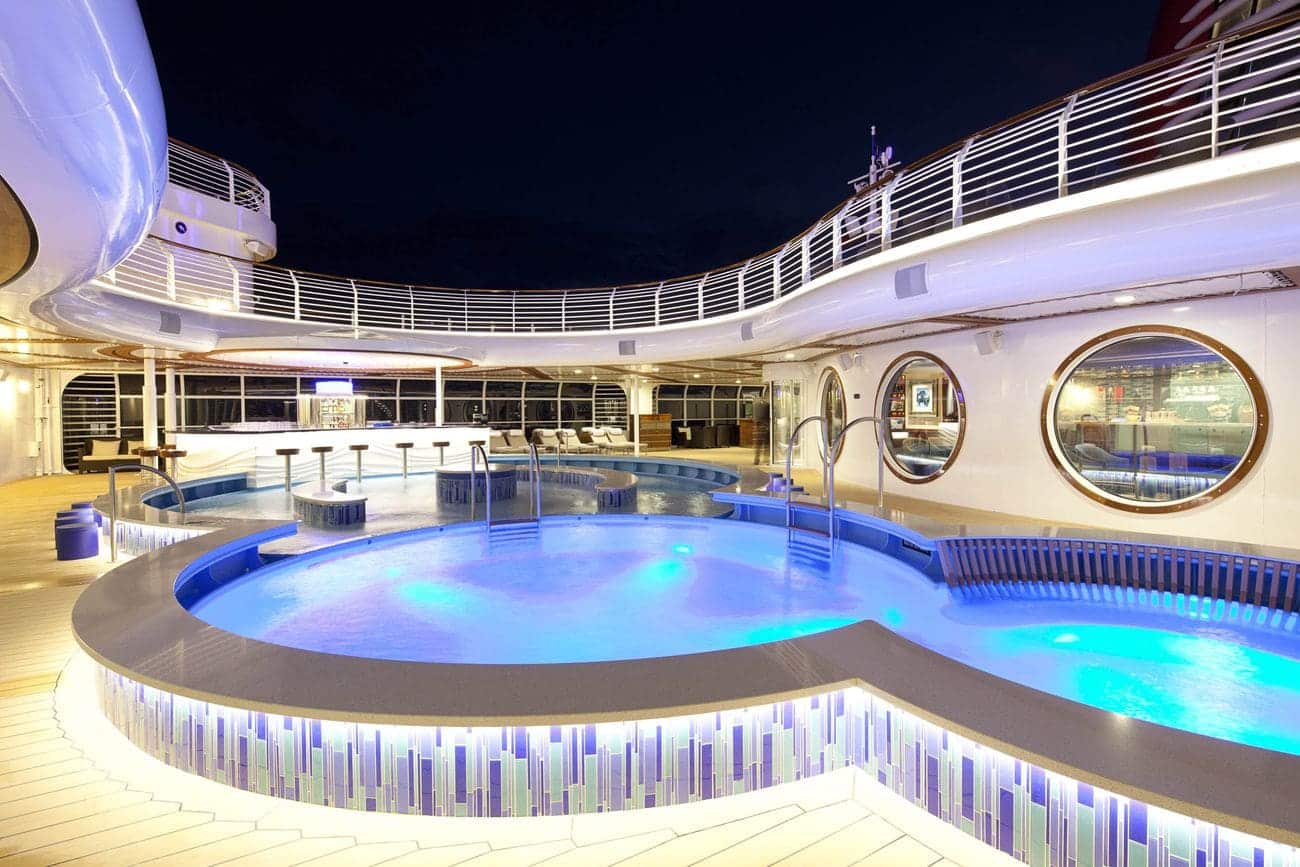 The Quiet Cove pool area on Disney Dream illuminated at night with glowing blue water curved seating and a nearby bar