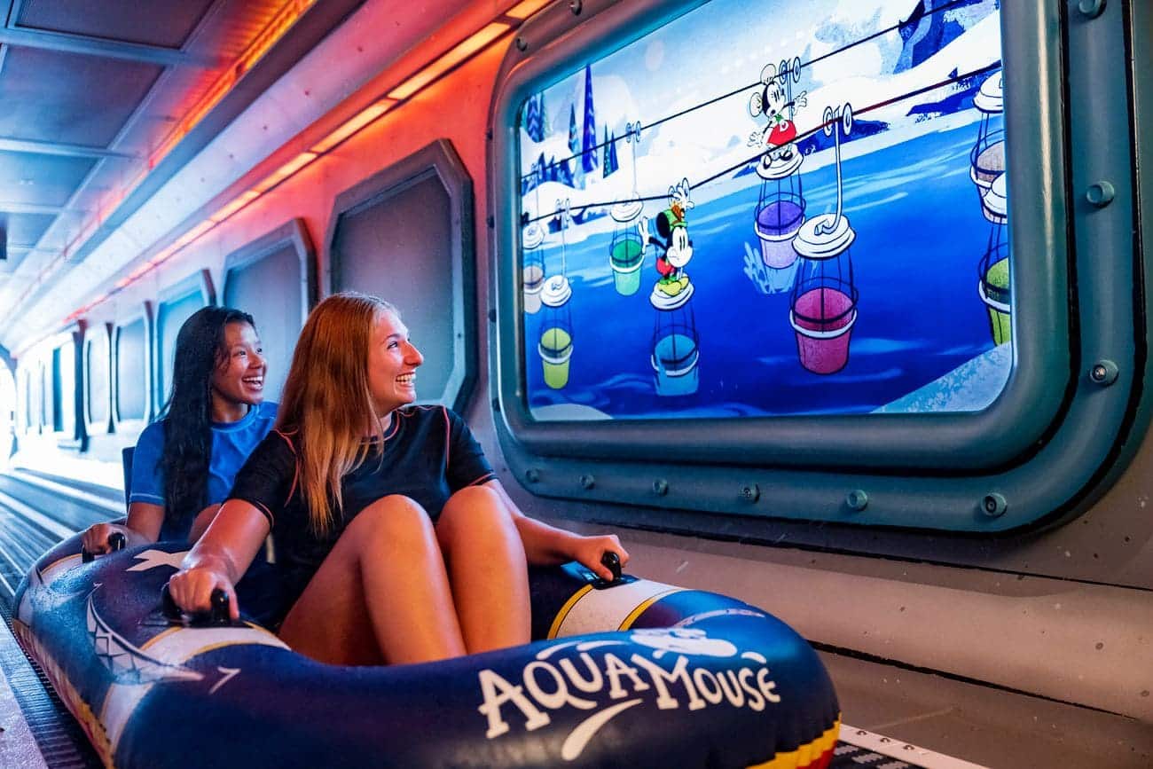 Guests riding the AquaMouse attraction on Disney Wish while passing animated Mickey Mouse scenes displayed inside the enclosed tunnel