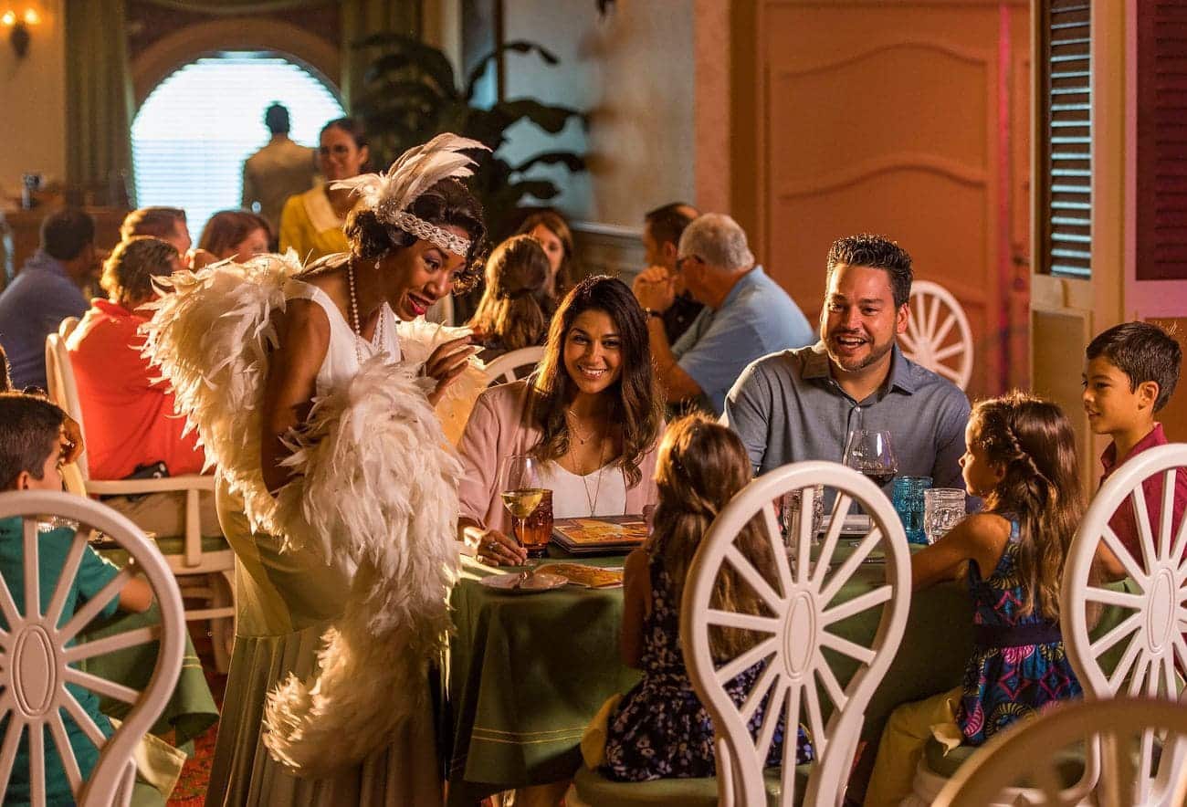 A family enjoying dinner at Tiana’s Place on Disney Wonder while a performer in feathered attire entertains guests