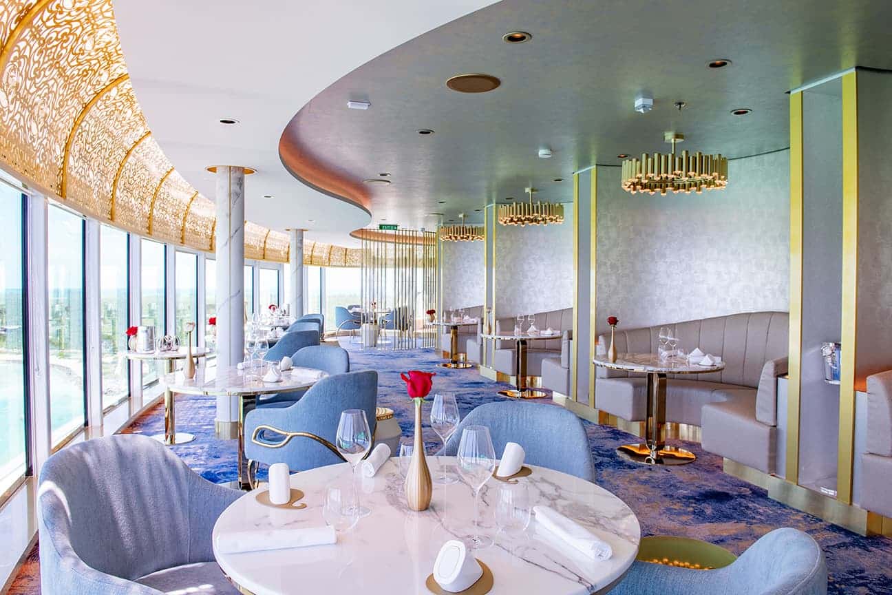The Enchanté restaurant aboard Disney Treasure featuring curved window walls ornate golden latticework and elegant marble dining tables