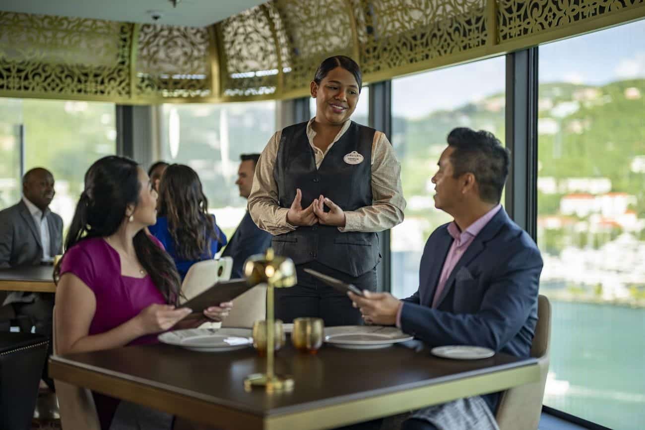 Guests dining at Palo Steakhouse with a server presenting menus in an elegant space lined with carved golden accents and large windows