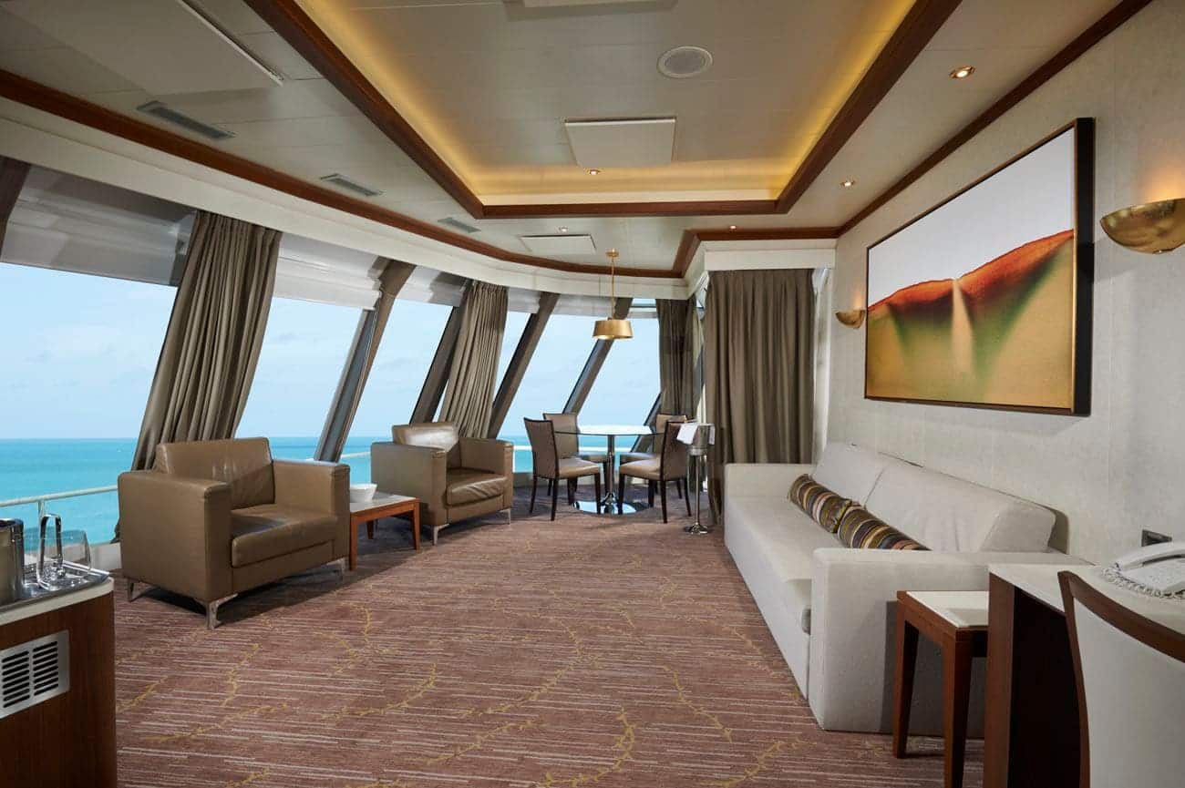 Deluxe Owner’s Suite aboard Norwegian Dawn featuring floor-to-ceiling windows, lounge seating, dining area, and panoramic ocean views