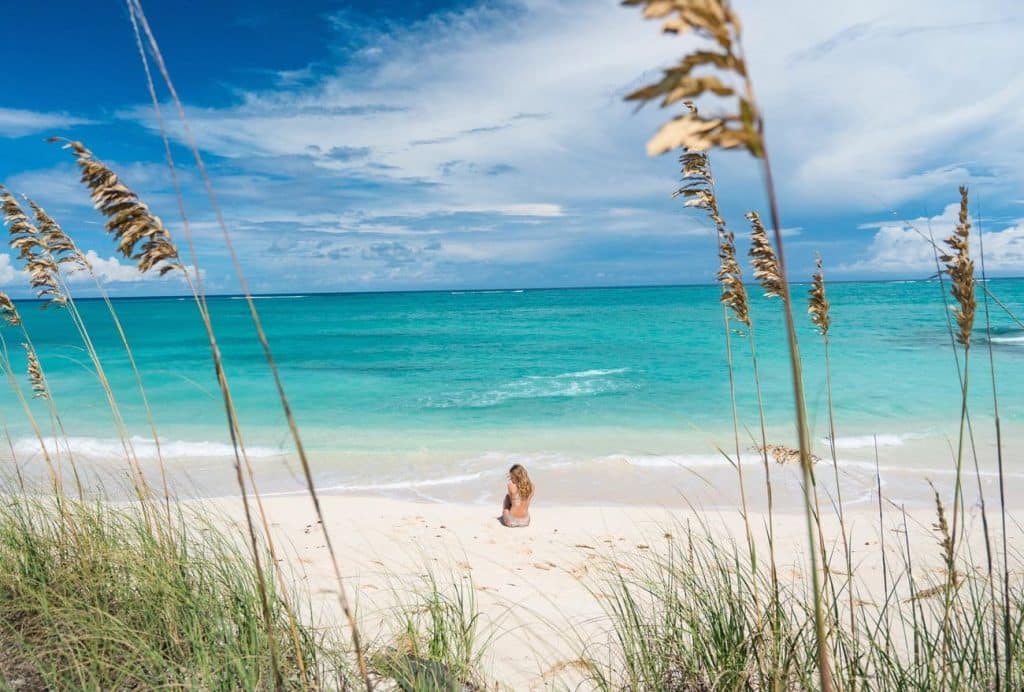 Woman sitting on a quiet white-sand beach in Nassau overlooking clear turquoise water under a partly cloudy sky