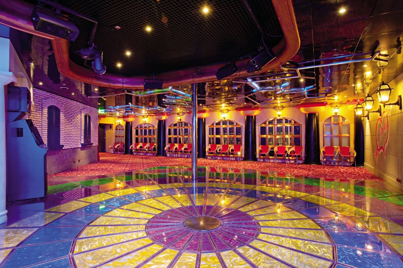 Colorful youth lounge with illuminated dance floor, mirrored ceiling panels, and rows of lounge chairs along decorative walls