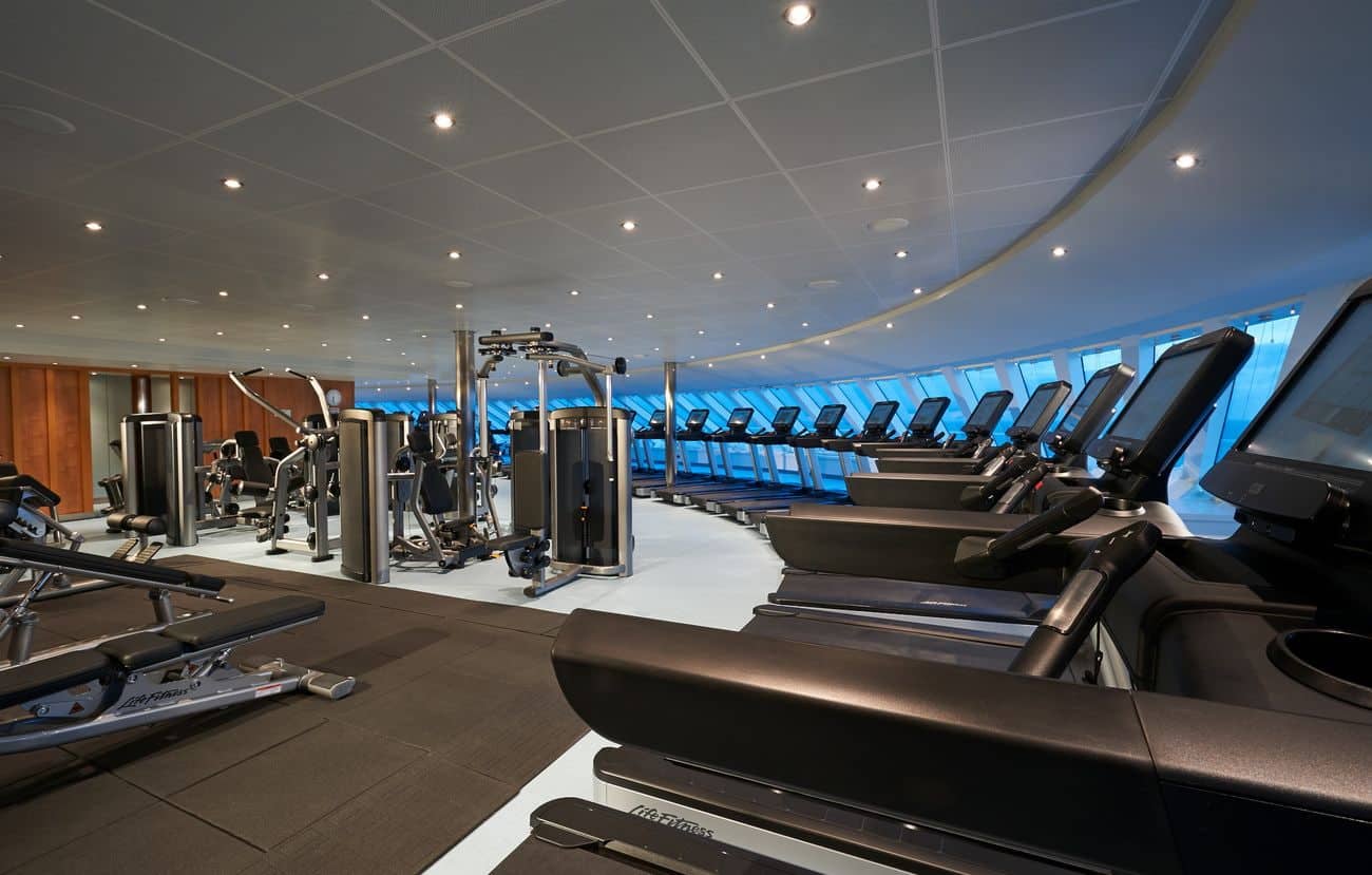 Ocean-view cruise ship gym with treadmills, weight machines, and wide forward-facing windows