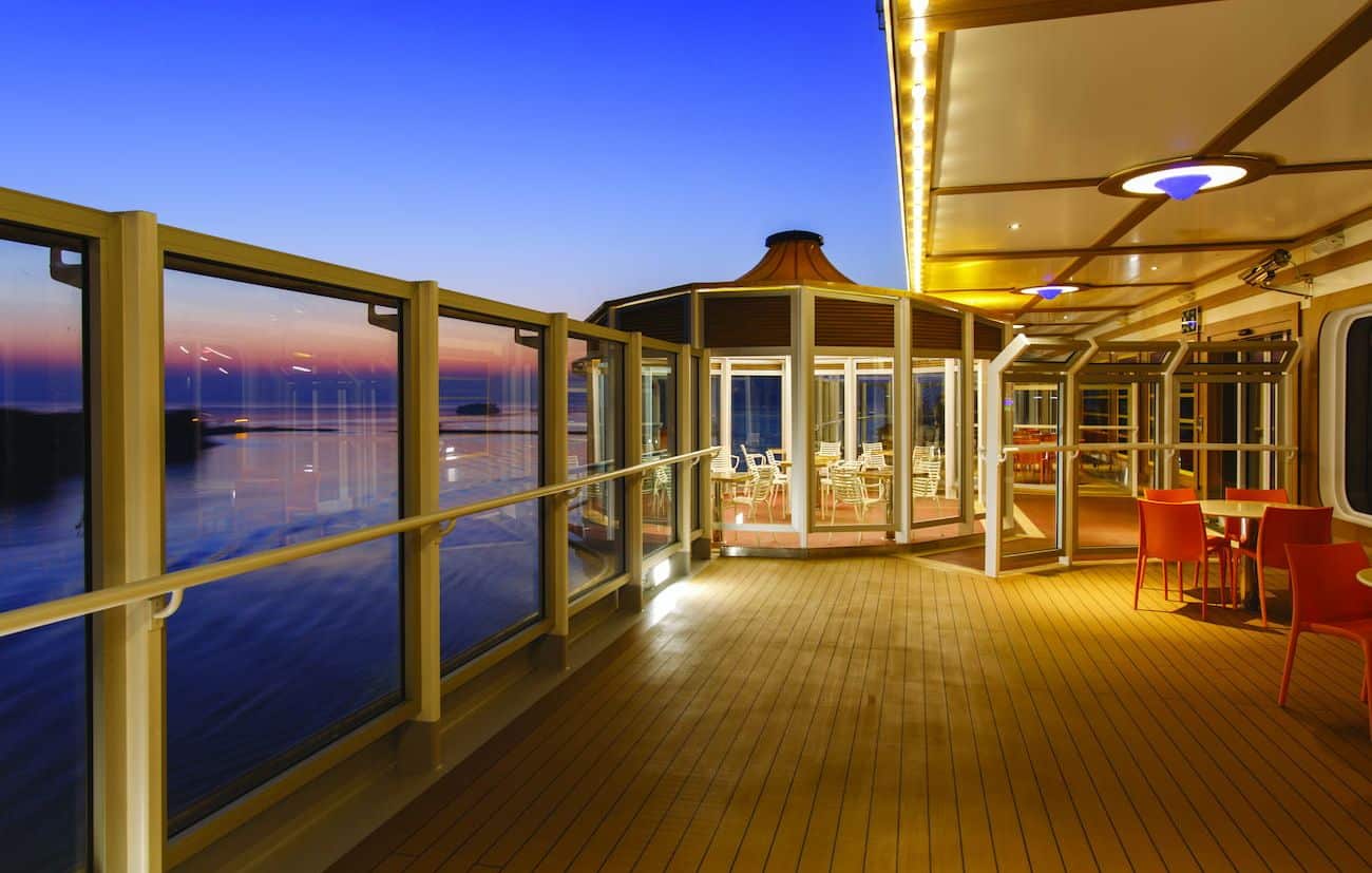 Costa Diadema open-air promenade at sunset with teak deck, glass windbreaks, and sea views.