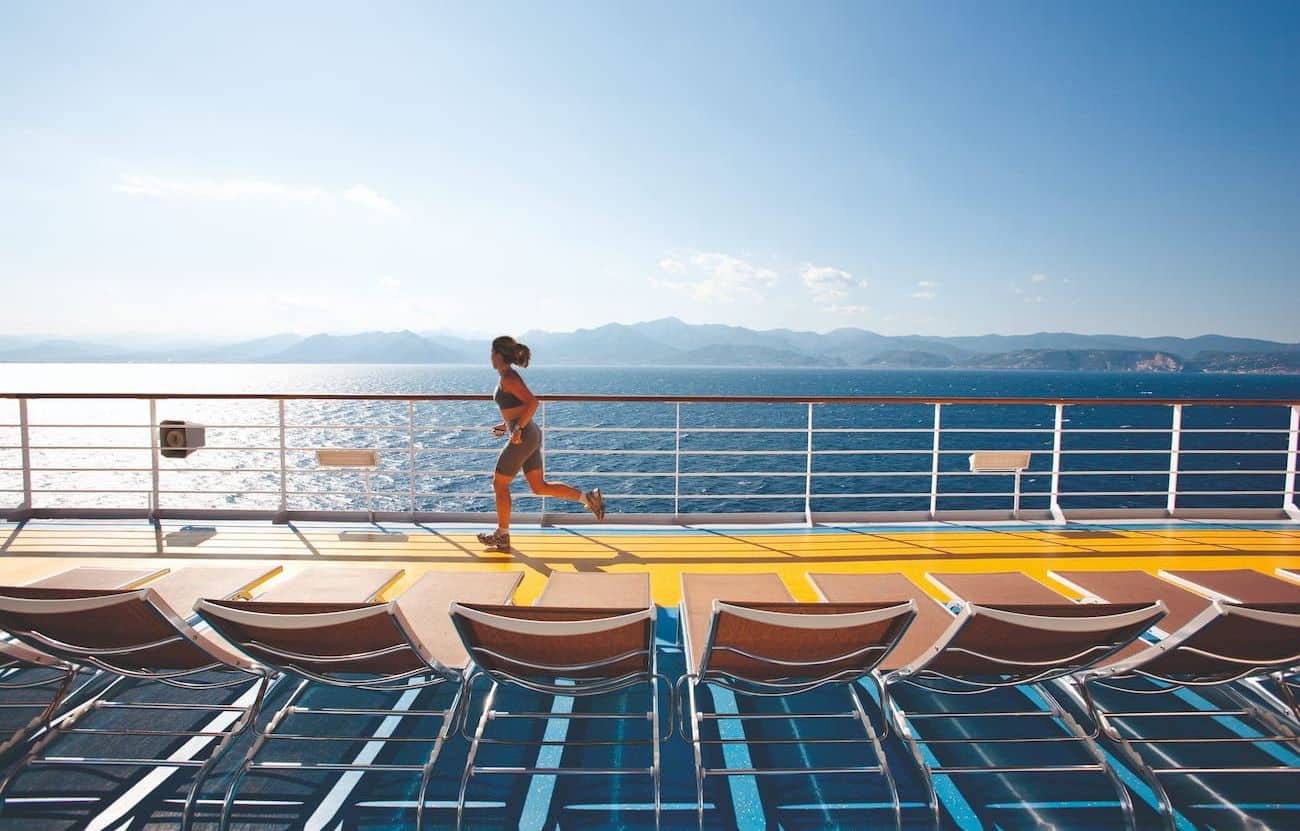 Costa Pacifica Open Deck jogging track with a runner, sun loungers, sparkling wake, and distant mountain views