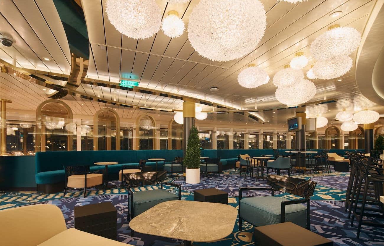 Kartel Bar lounge with glowing globe lights, teal banquettes, patterned carpet, and small tables arranged across a wide interior space.