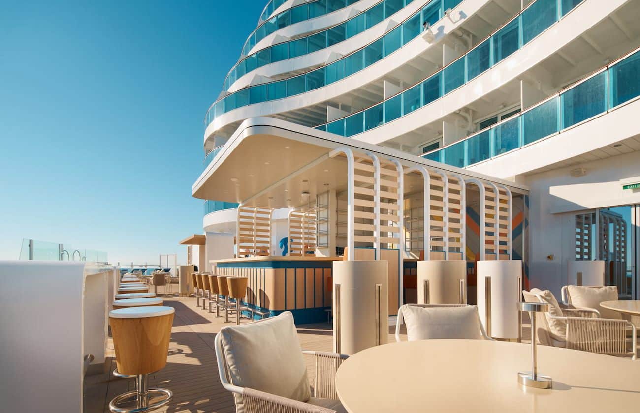 Costa Toscana Terrazza Infinity with outdoor seating, bar stools, pale furnishings, and a bright sea-facing deck under clear skies.