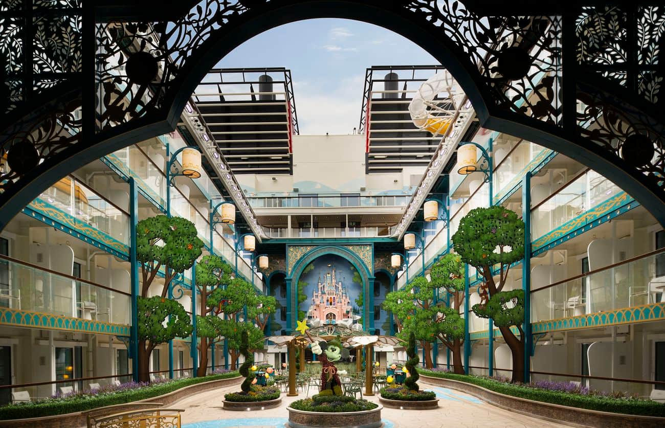 Disney Imagination Garden courtyard framed by ornate arches, with sculpted greenery, balconies, and a fairy-tale castle mural